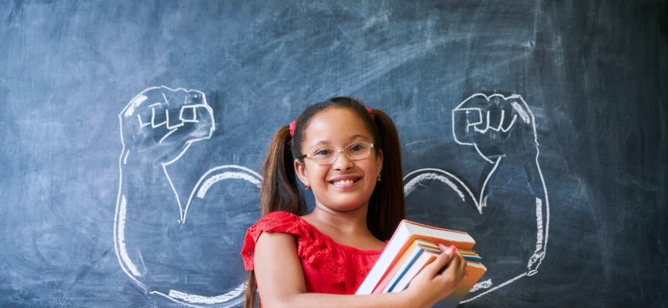 Concept on blackboard at school. Intelligent and successful hispanic girl in class. Portrait of female child smiling, looking at camera, holding books against drawing of muscles on blackboard